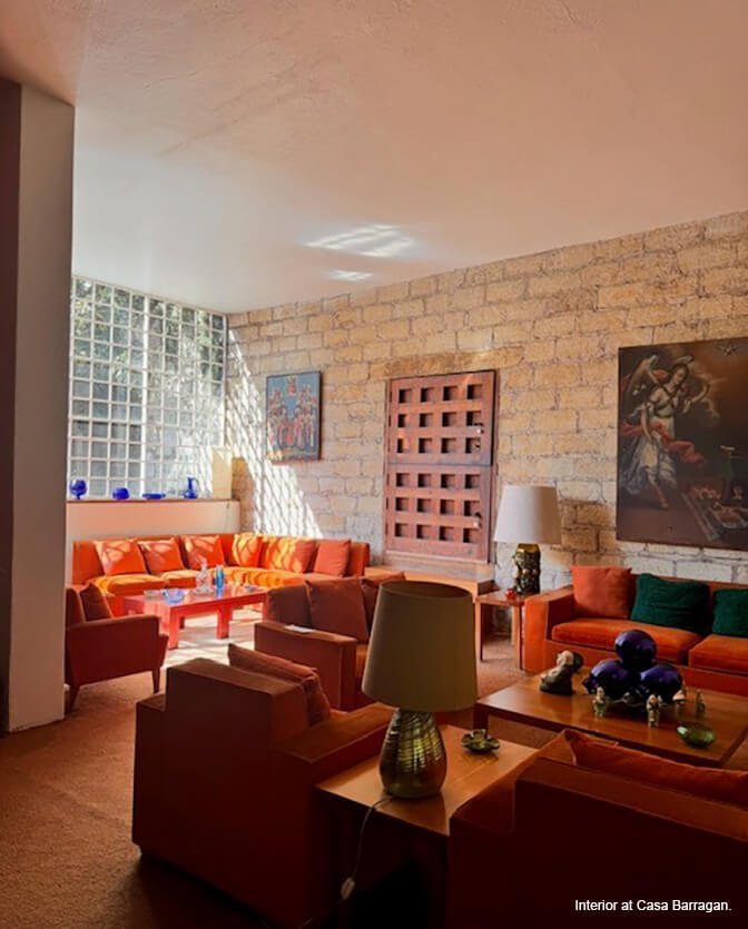 An interior with a glass grid window, a pale brick back wall, a geometric wooden door and bright orange sofas and chairs.