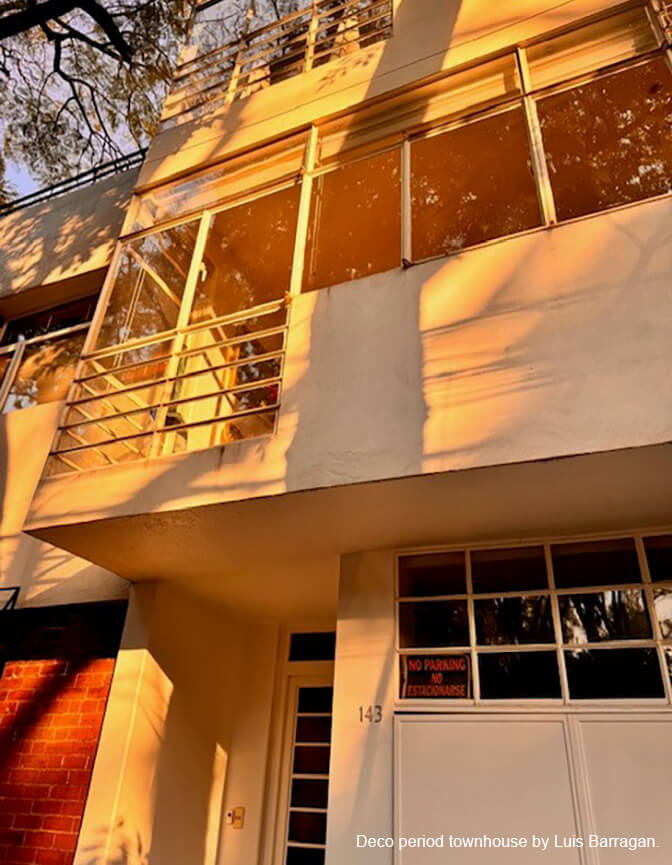 A white deco style facade with window and railings and red brick in the lower corner with a tint of yellow sunshine.