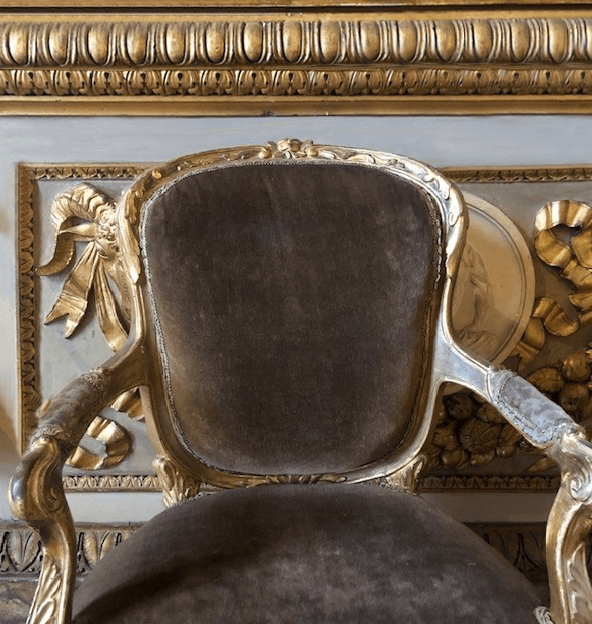 A antique chair with ornate carving and gilding and muted brown velvet back and seat cushions against a decorative white wall with gilding.