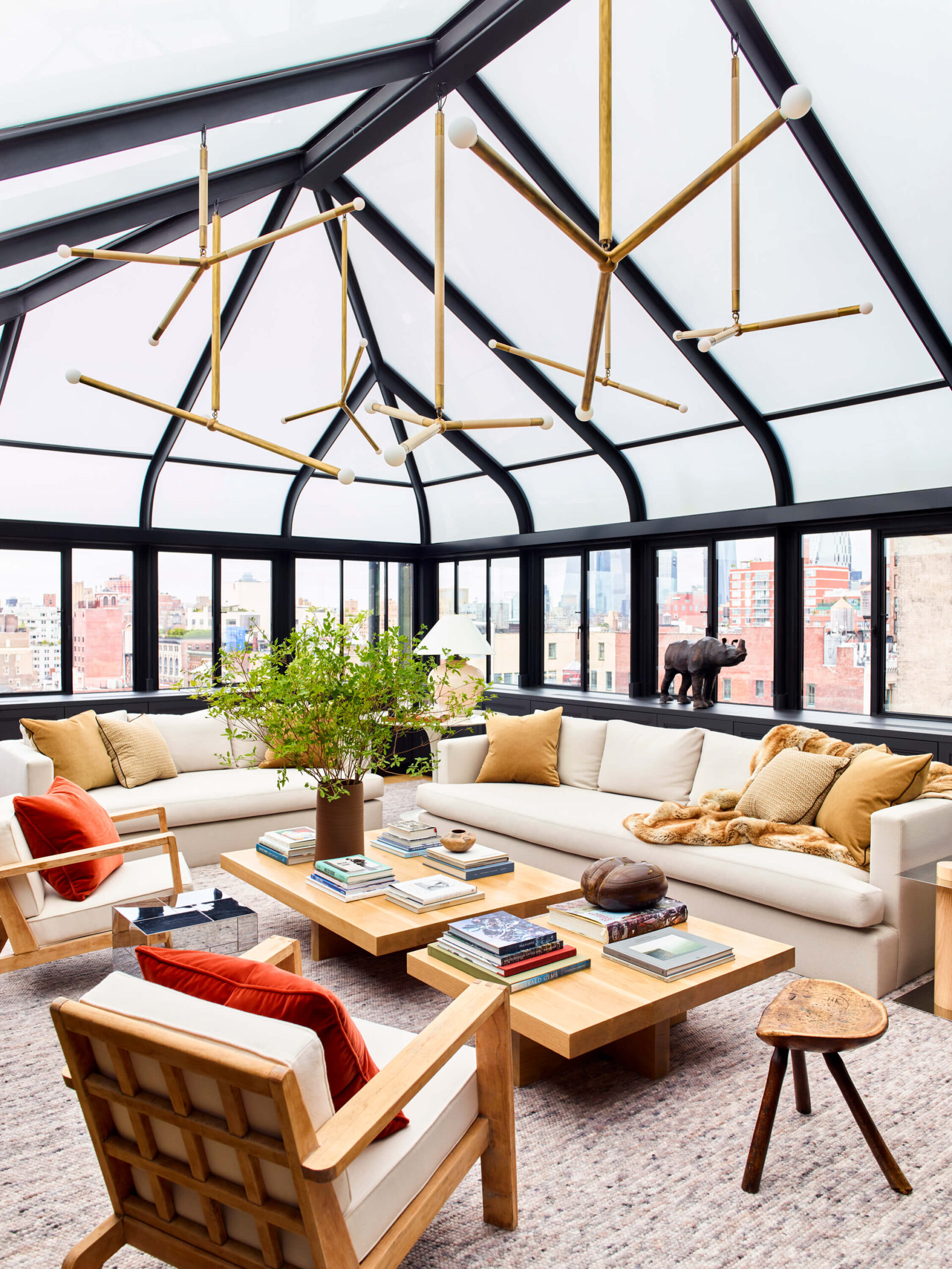 A greenhouse living room with an abstract light fixture, neutral rug, beige sofas, wood club chairs and wooden coffee tables.