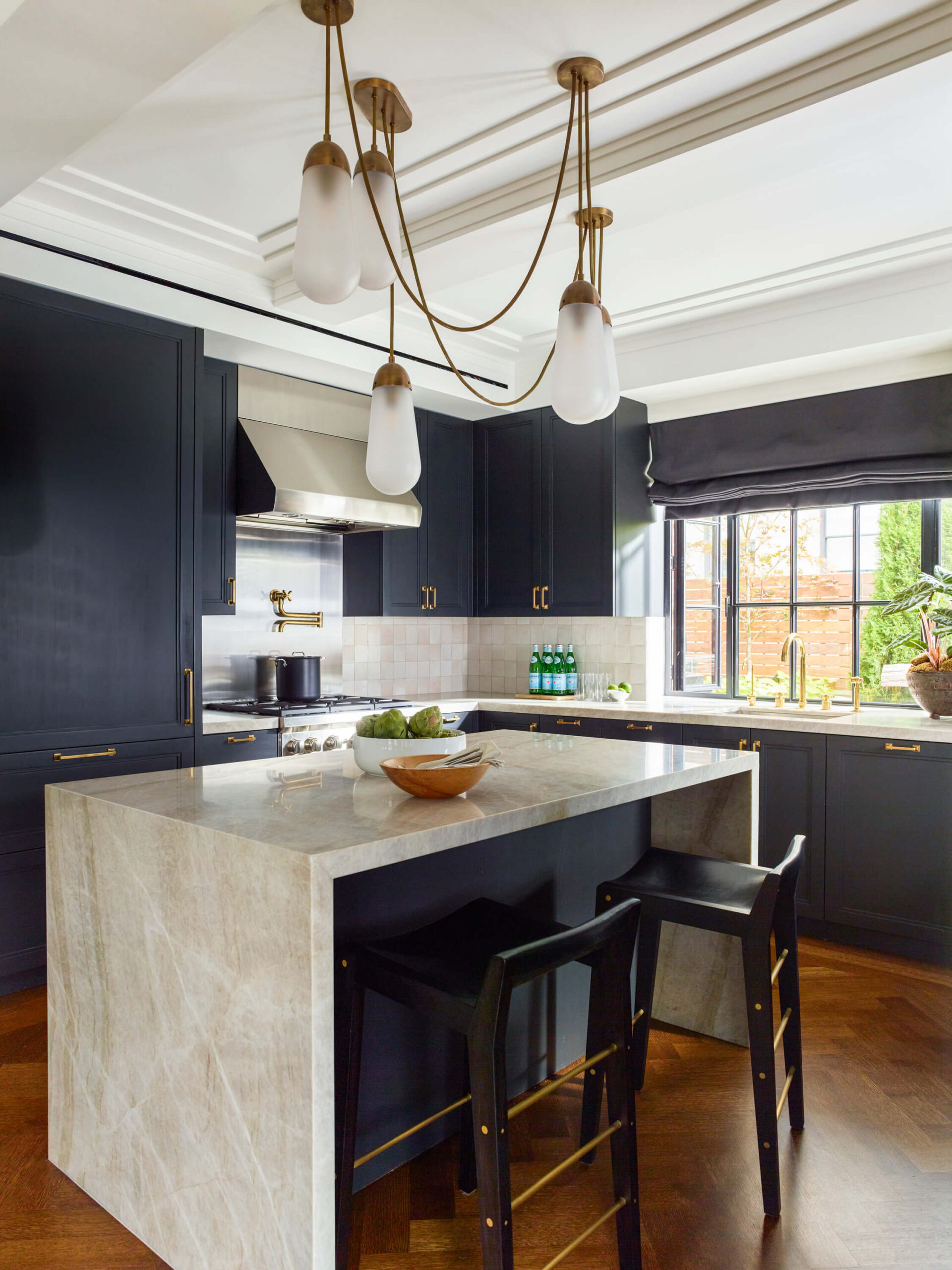 Modern kitchen with black cabinetry and beige tile walls, a beige stone island with black stools and a five-light pendant.