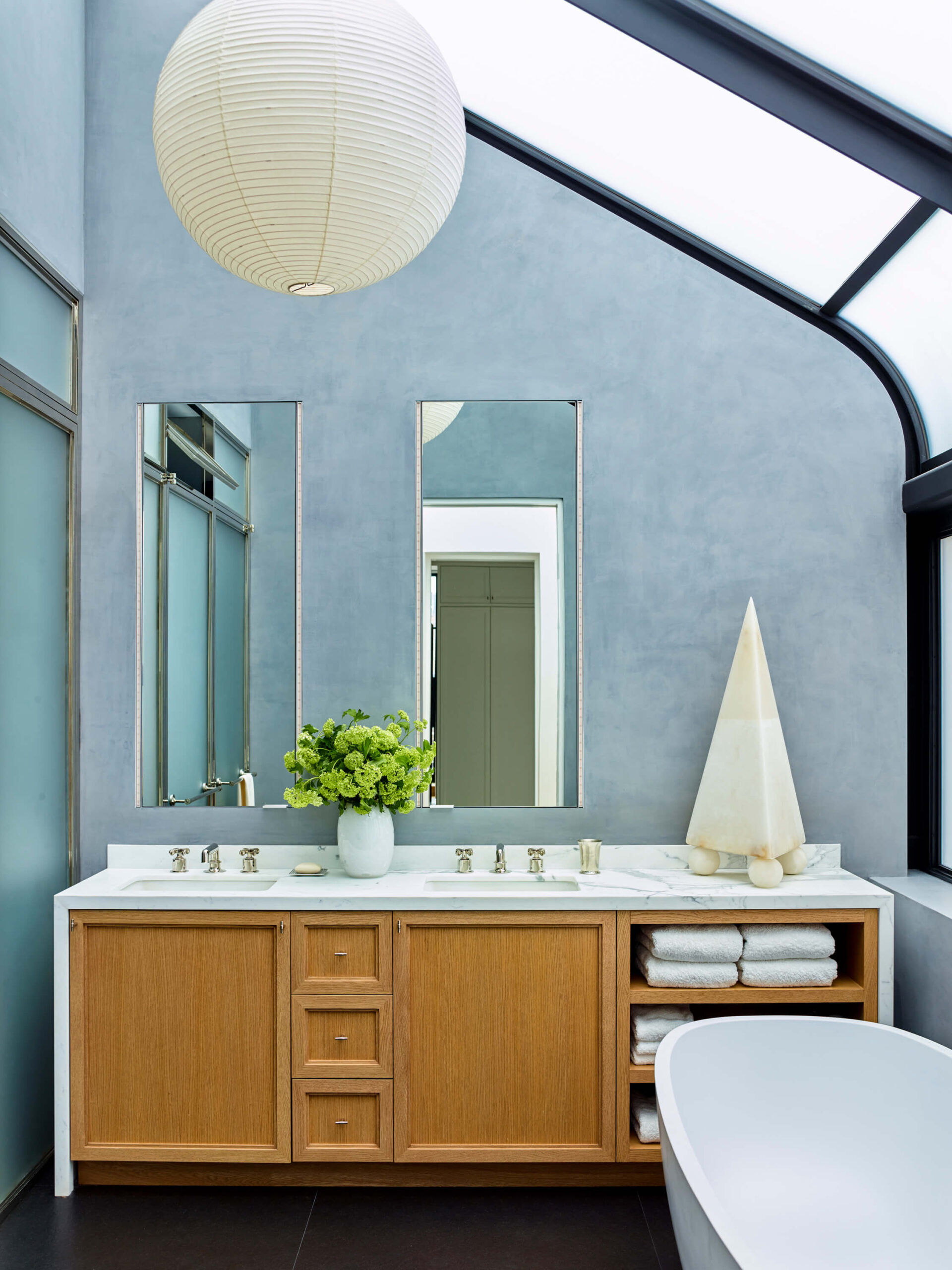 A double sink bathroom vanity in blond wood an a white top with two vertical mirrors a lantern pendant and a light blue wall.