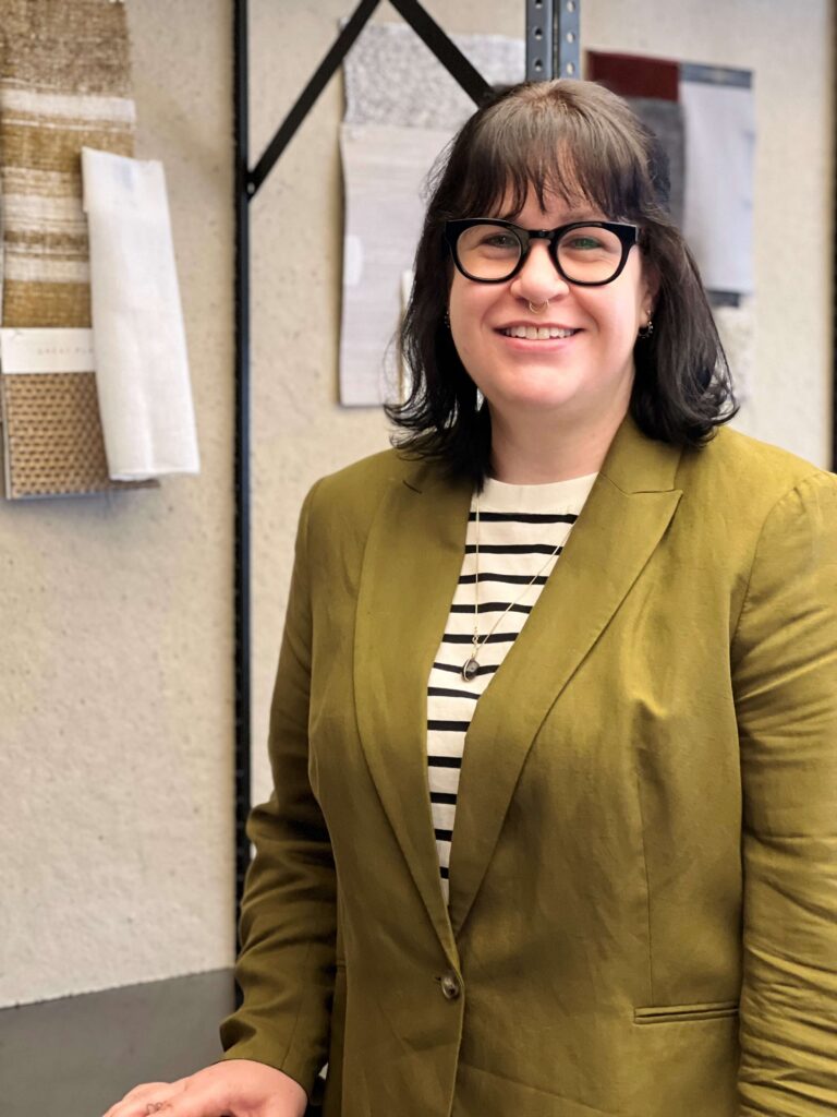 A dark-haired woman with glasses and a chartreuse blazer on standing in front of a wall pinned with fabric samples.