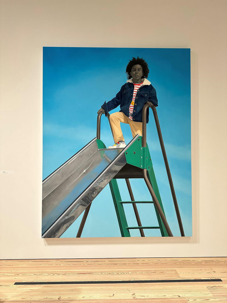 A photo-realistic painting of a boy in yellow pants and a denim jacket, standing at the top of a green slide. entitled Kingdom, by Amy Sherald.