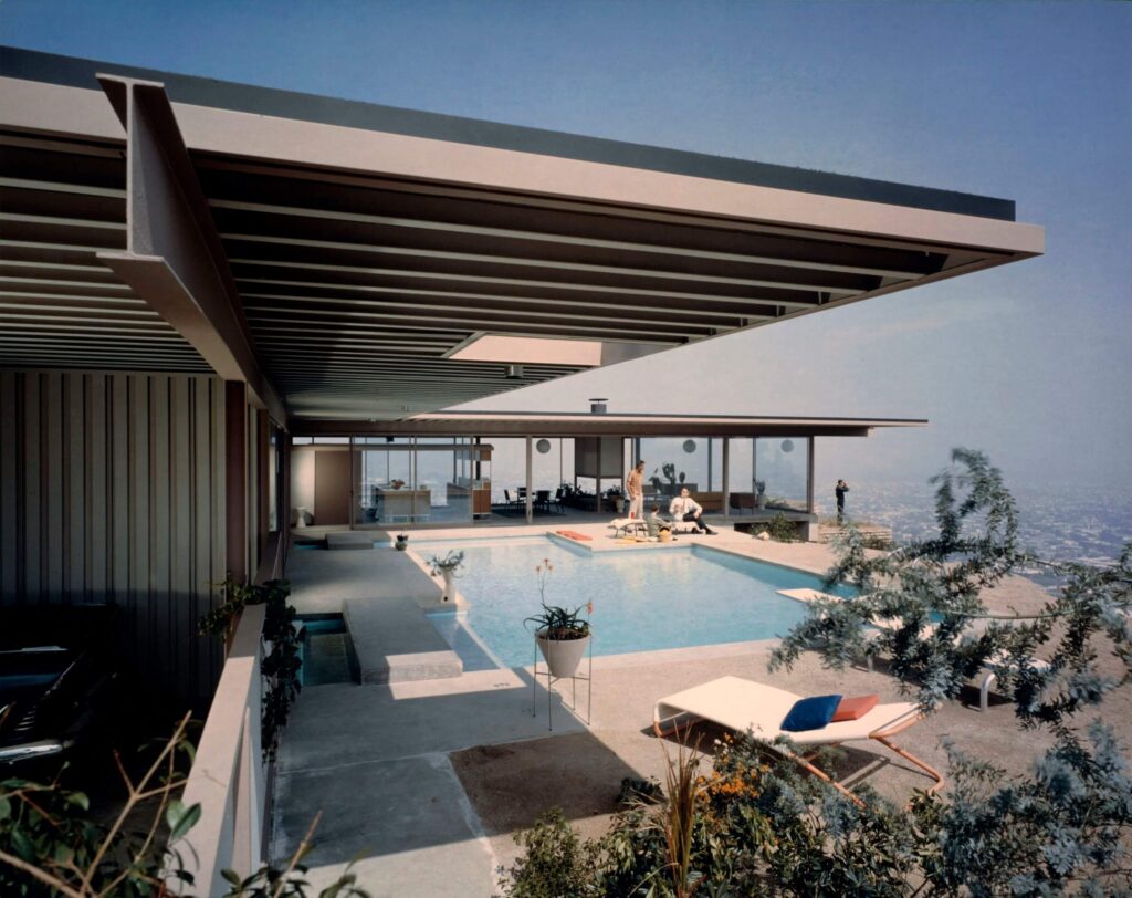 A modern house with a cantilevered roof extending over a concrete pool deck with plants and lounge chairs overlooking LA.
