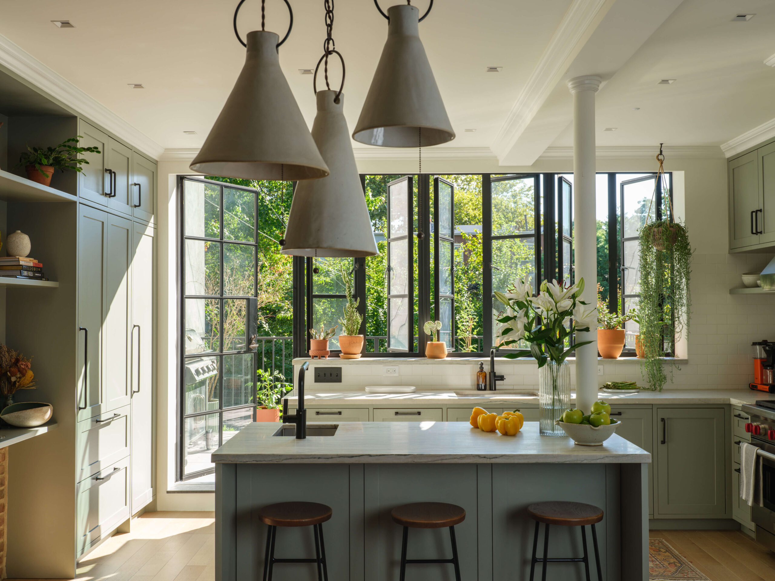 Airy kitchen with back wall of casement windows and doors. Gray cabinets with stone counters. Three ceramic pendant lights.