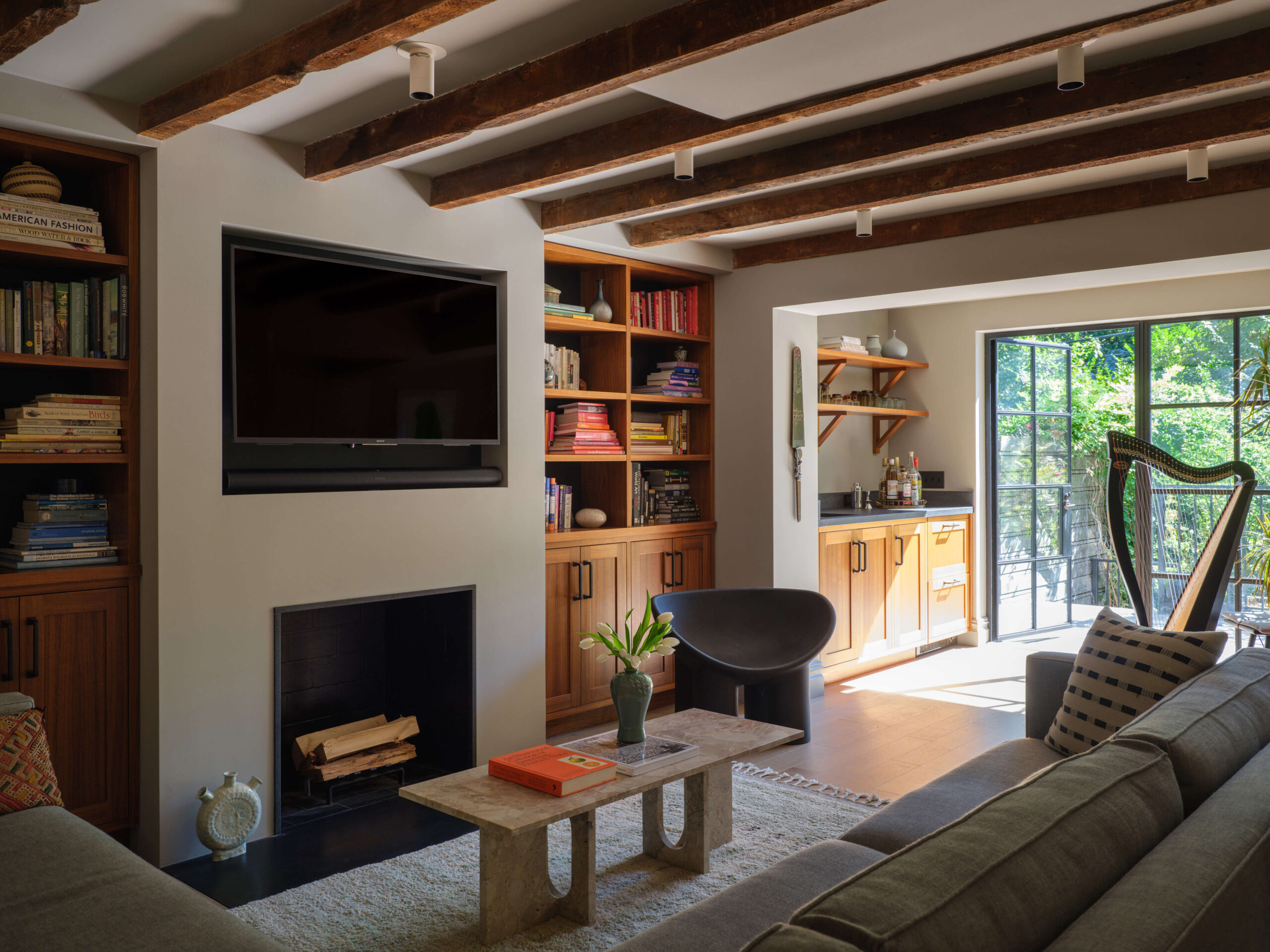 Den with white walls, exposed wood ceiling beams, black inset fireplace and tv. Built-in shelving sofa and coffee table.
