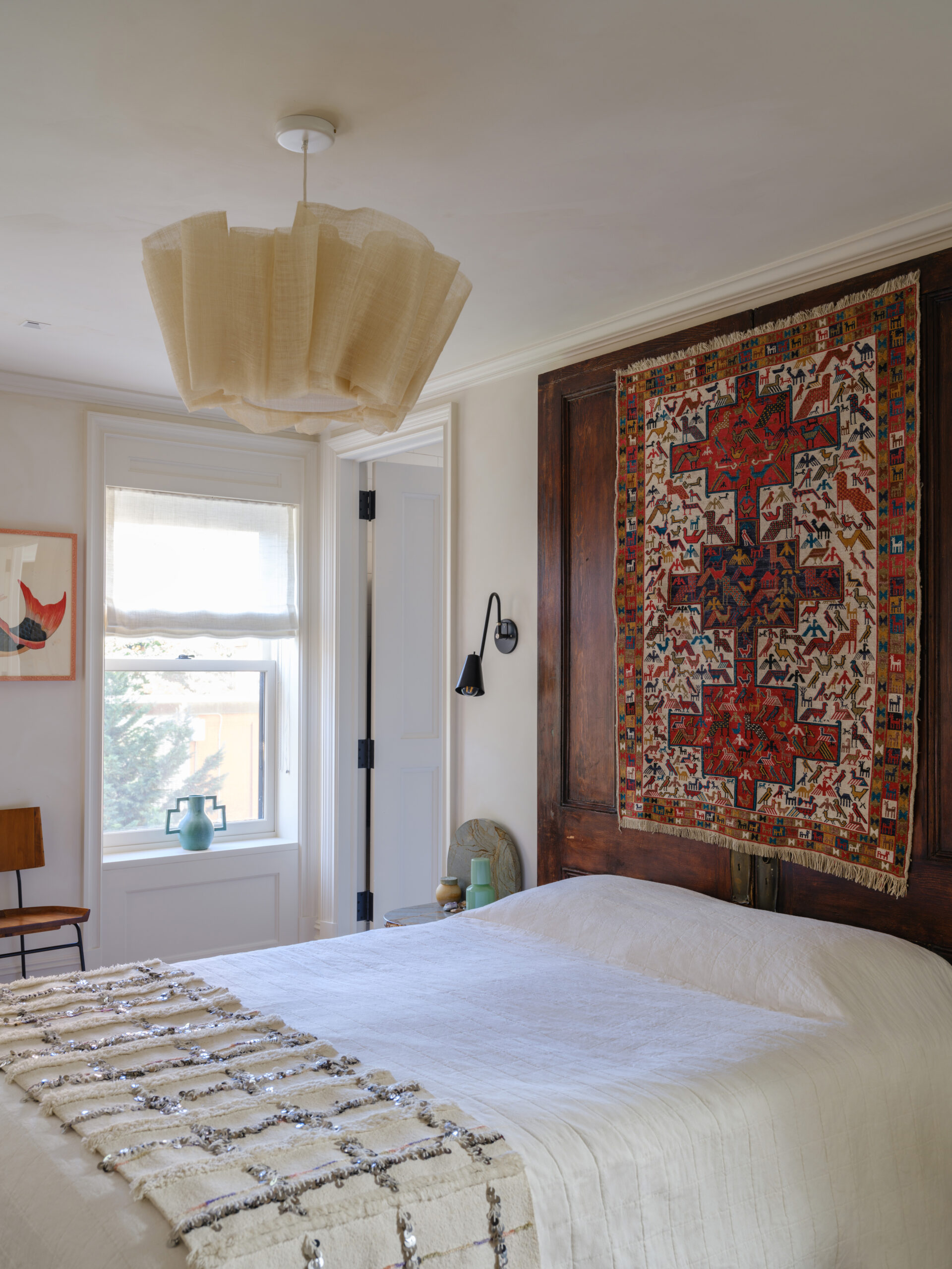 White bedroom with a pair of tall wood headboard and tapestry, a window with a sheer white shade and beige ceiling light.