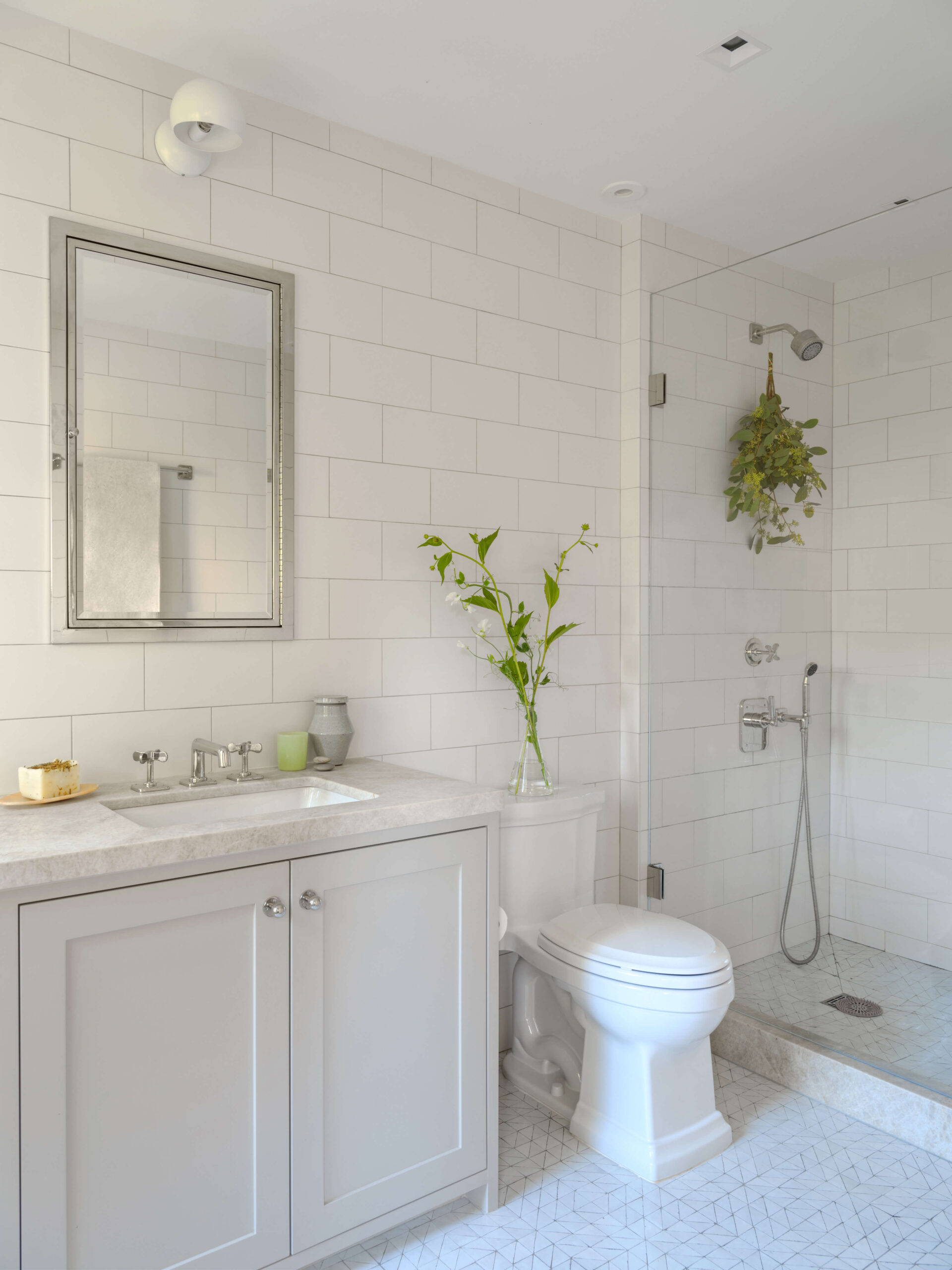 All-white bathroom with subway tiles, and tile floor, sink vanity and mirror, toilet and standing shower. Greenery as décor.