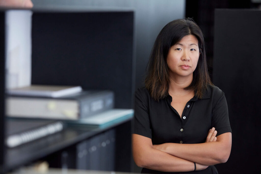 A woman in a black top, with arms crossed, standing amidst shelving with some books in the foreground.