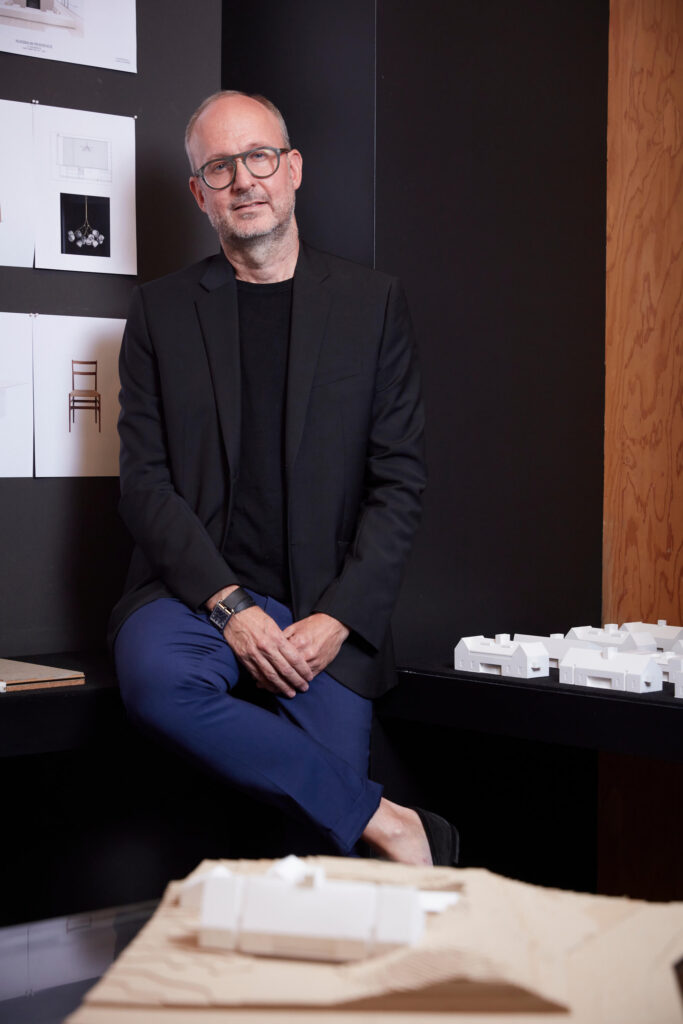 A man in a black shirt and jacket with dark blue pants, sitting in a design studio with architectural drawings and models.