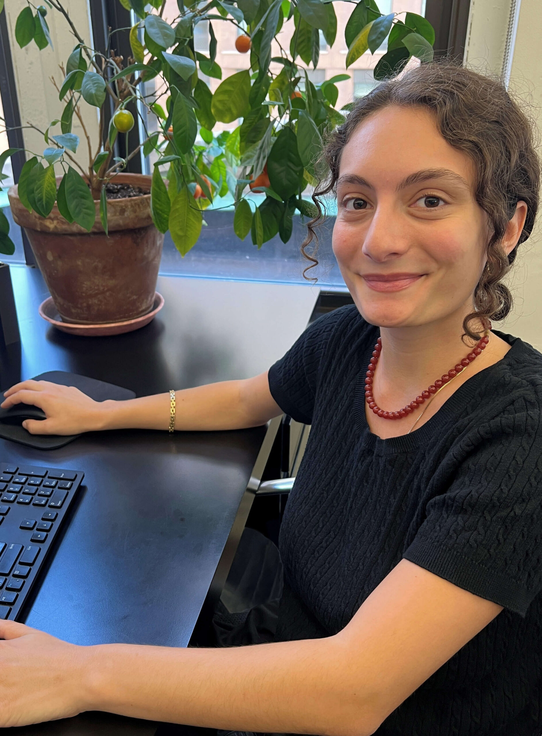 A young woman with brown curly hair, in a black sweater, sitting at a black desk with a plant.