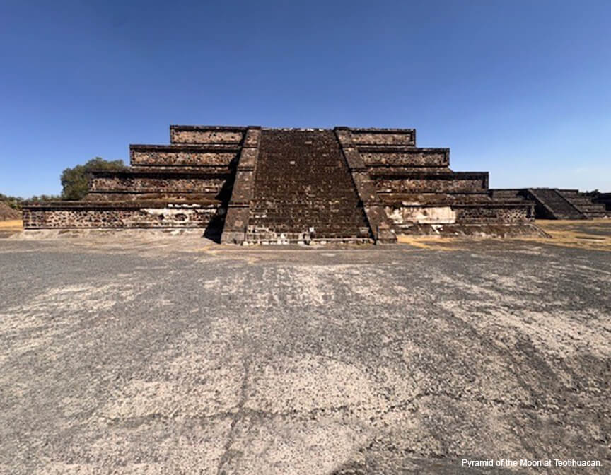 A dark stone monument with flat levels and a central stair; a blue sky in the background and asphalt in the foreground.