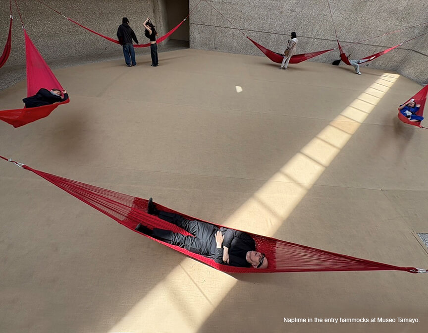 An open space with a neutral floor, concrete walls and an installation of large red rope hammocks for visitors to relax.