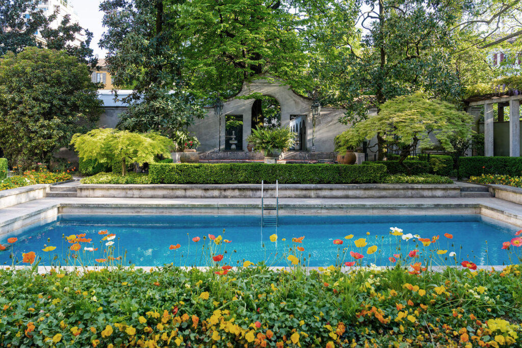 A rectangular pool with a stone deck; a garden of red, orange and yellow flowers in the foreground; trees in the background.