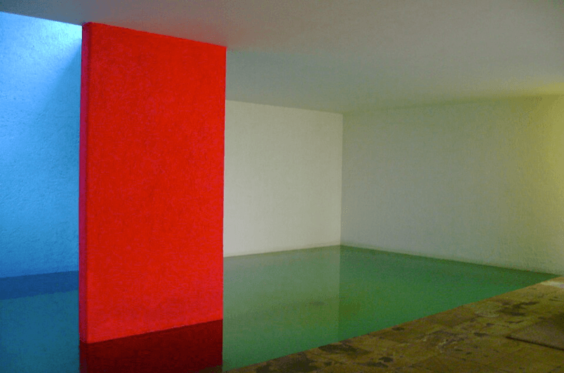 A view of Casa Gilardi's minimalist swimming pool enclosed in a white-walled room with a red wall in middle and blue light from the sky on the left.