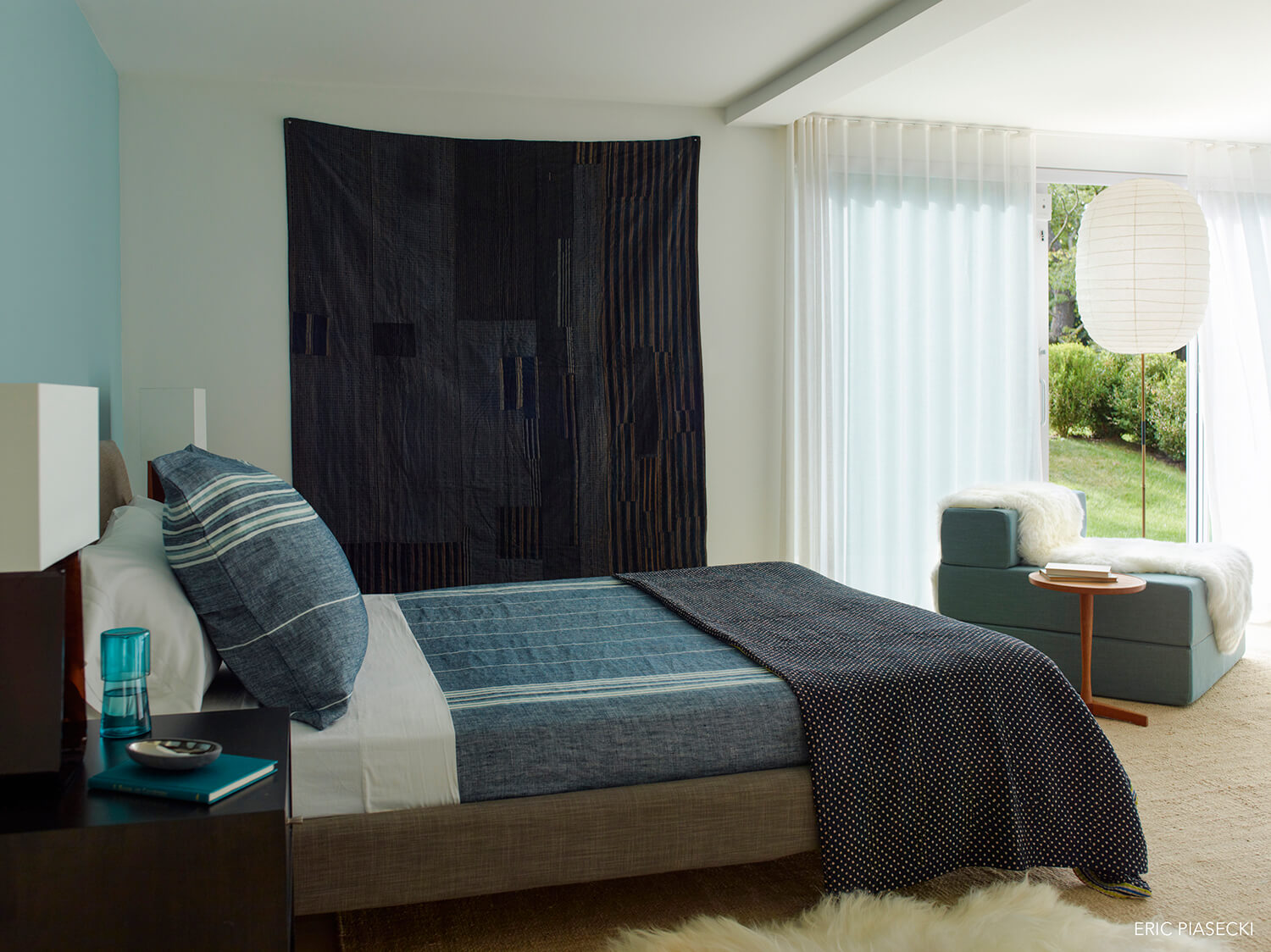 White bedroom, a sliding door to yard, sheer white curtains, a navy-blue wall-hanging and a bed with white and blue bedding.