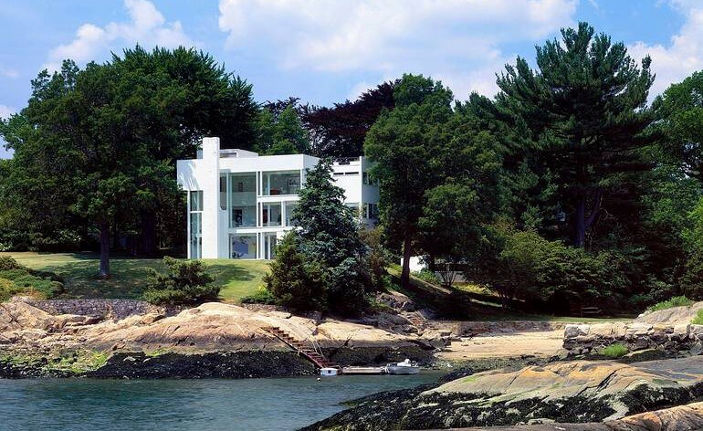 A white multistory, modern house nestled amongst trees. Rocks and water in foreground.