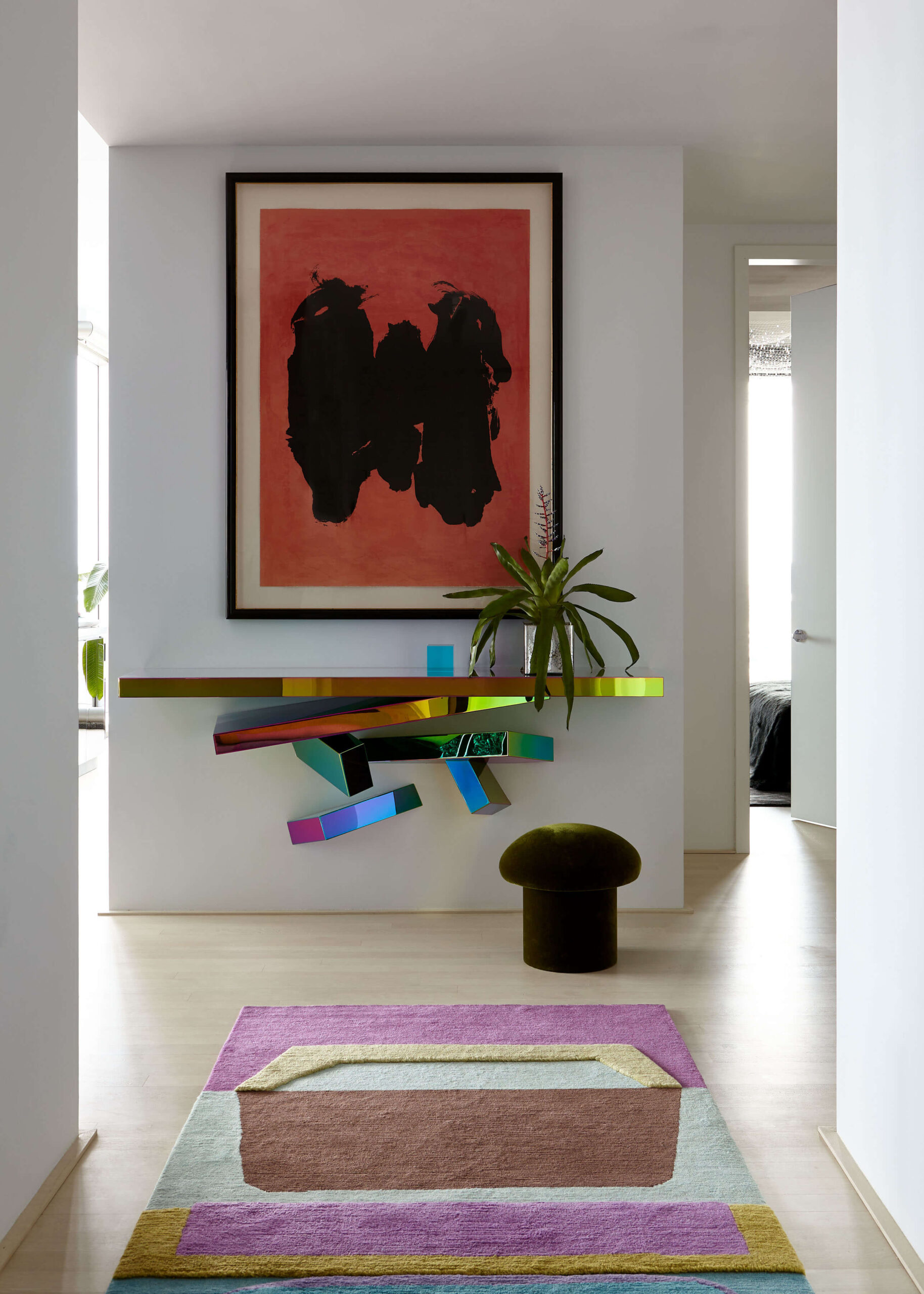 Hallway with white walls, red and black framed art, multi-colored metallic console table with plant and multi-colored rug.
