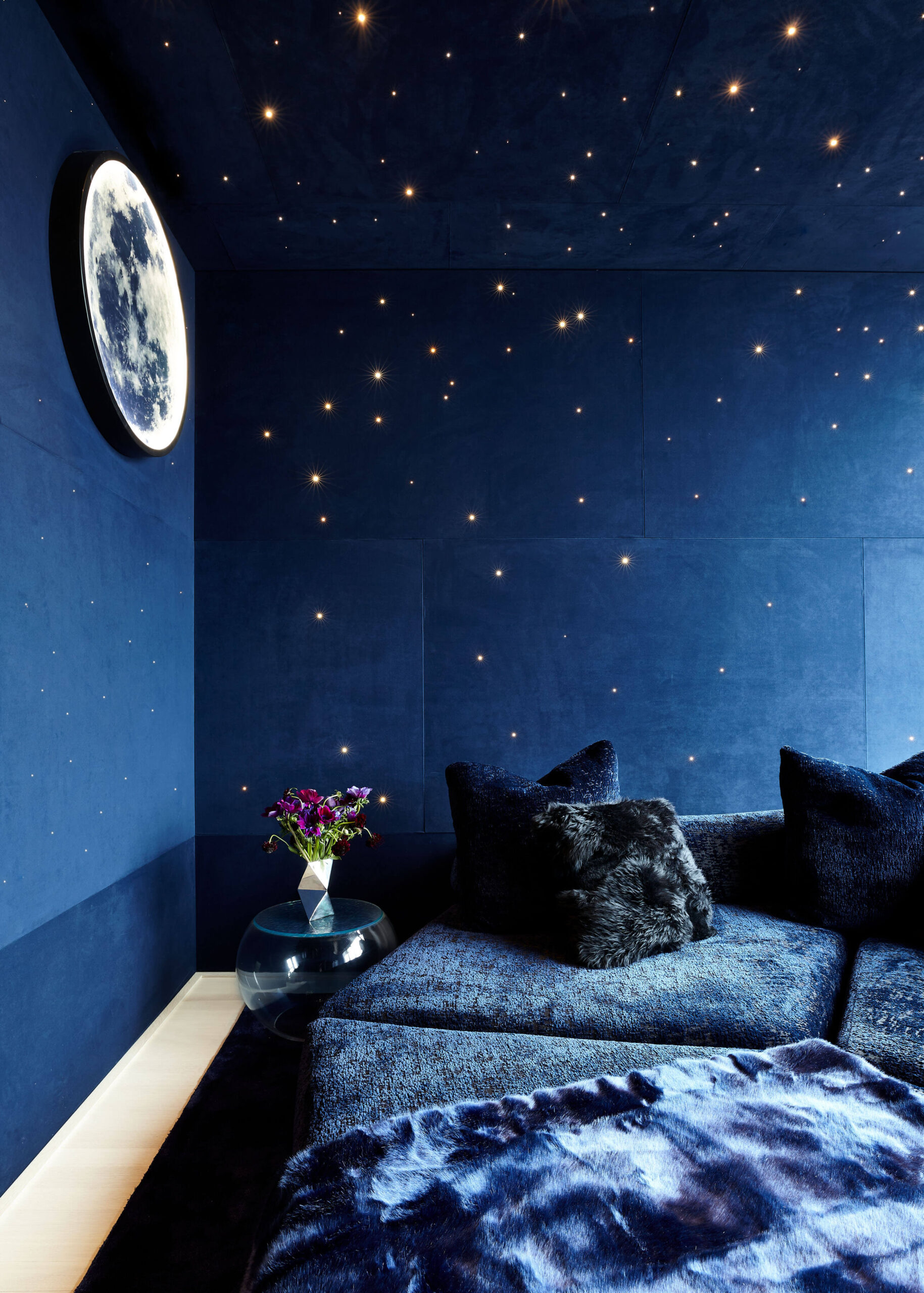 Media room with dark blue walls with a moon wall-hanging and constellation-inspired fiberoptic lighting and a coordinating blue upholstered sofa.