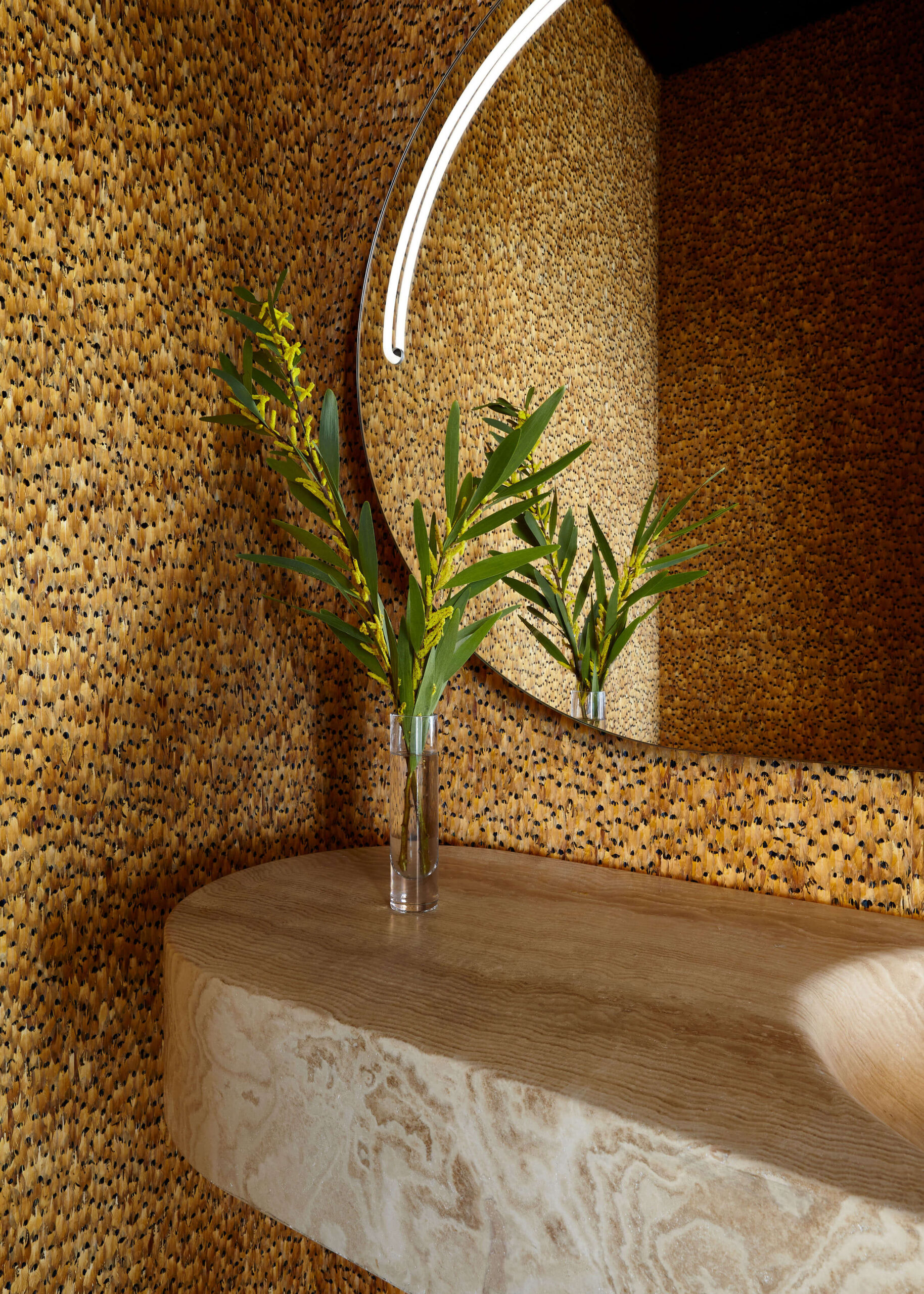 Powder room with round mirror, golden-colored feather wall covering and a sink vanity made of a beige marble block.