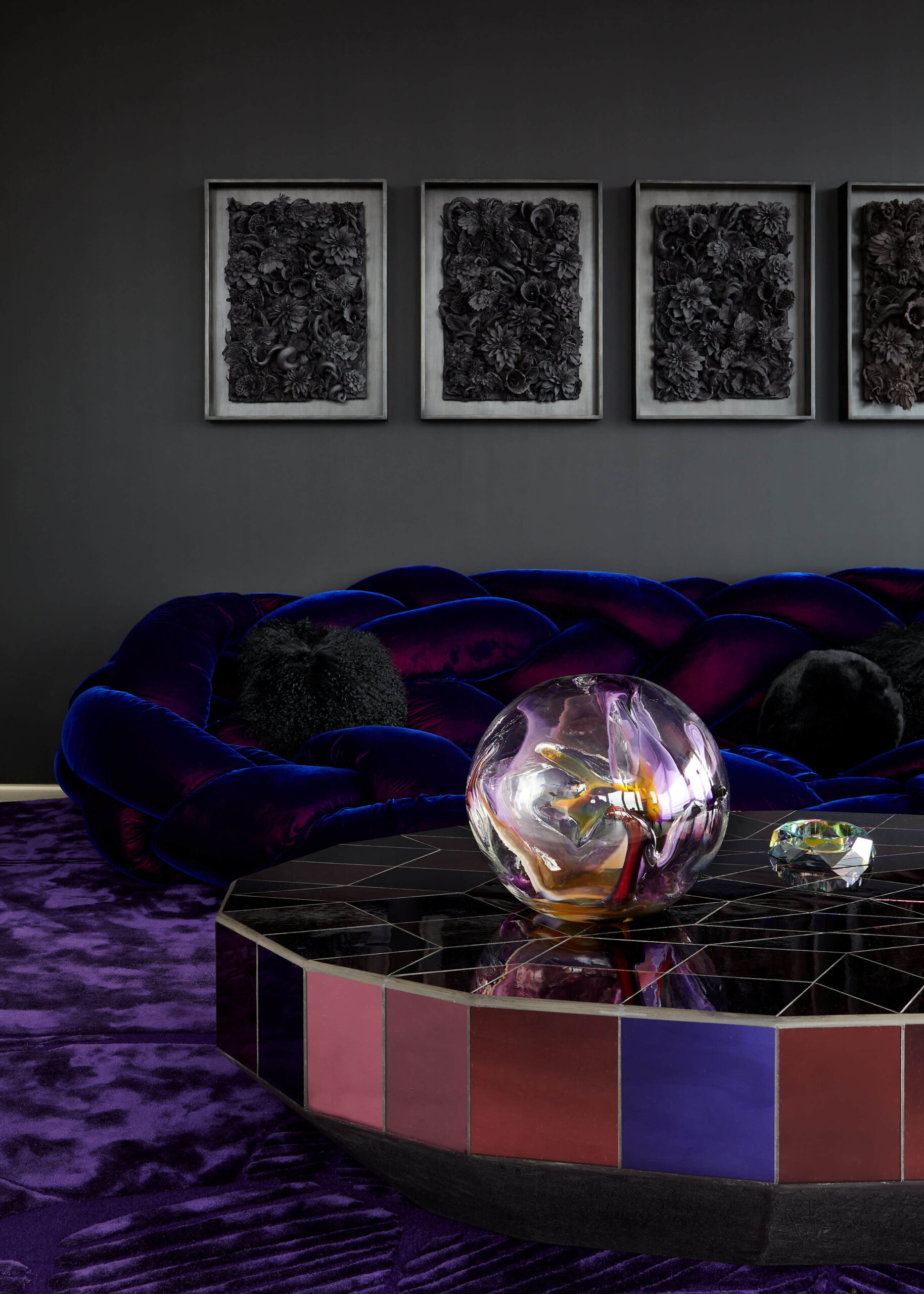 Living room with black wall, four framed art works, deep blue braided velvet sofa, purple carpet and abstract coffee table.