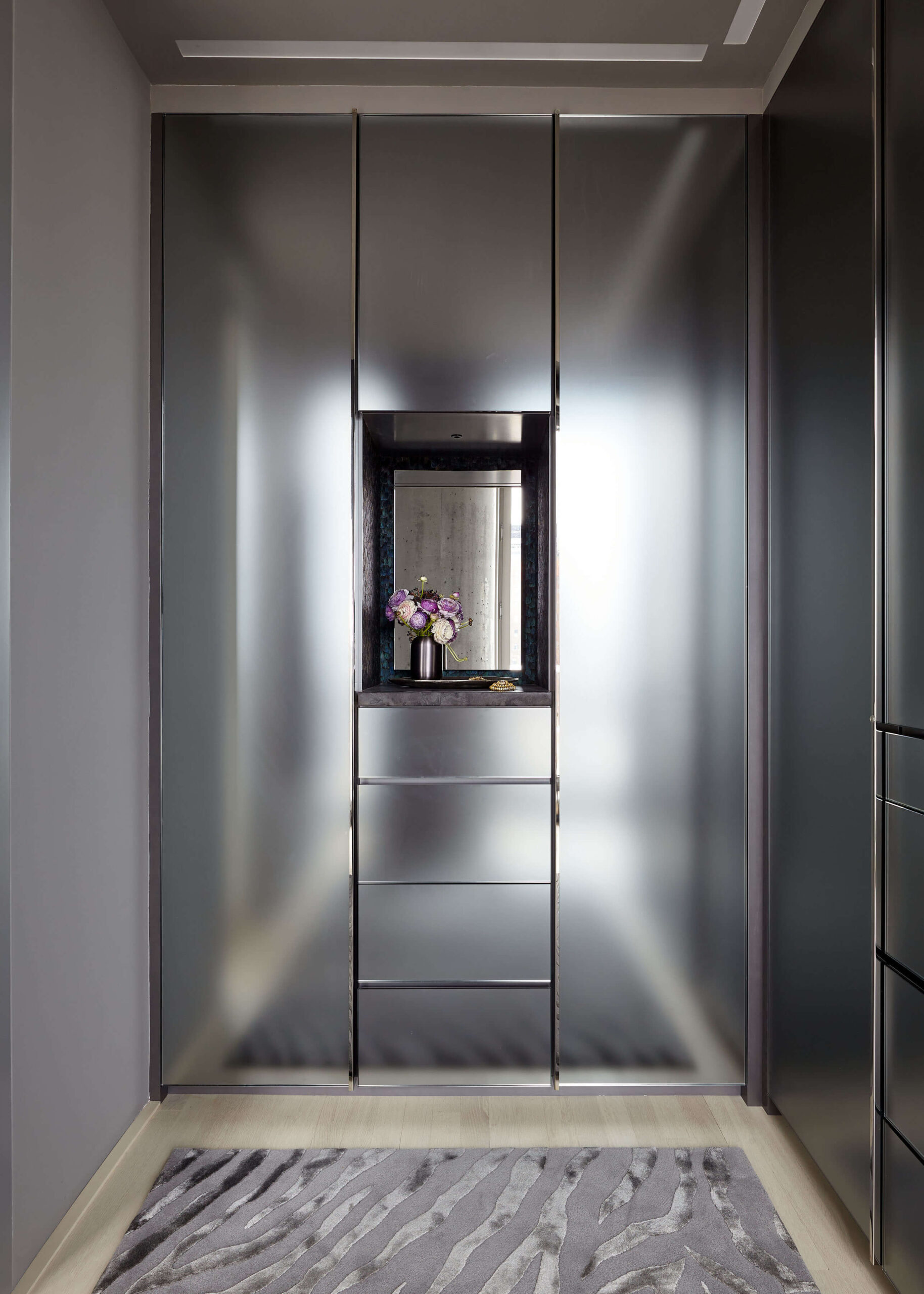 Walk in closet with cabinetry clad in dark gray acid-etched mirror, niche with vase of flowers in center, and gray rug.