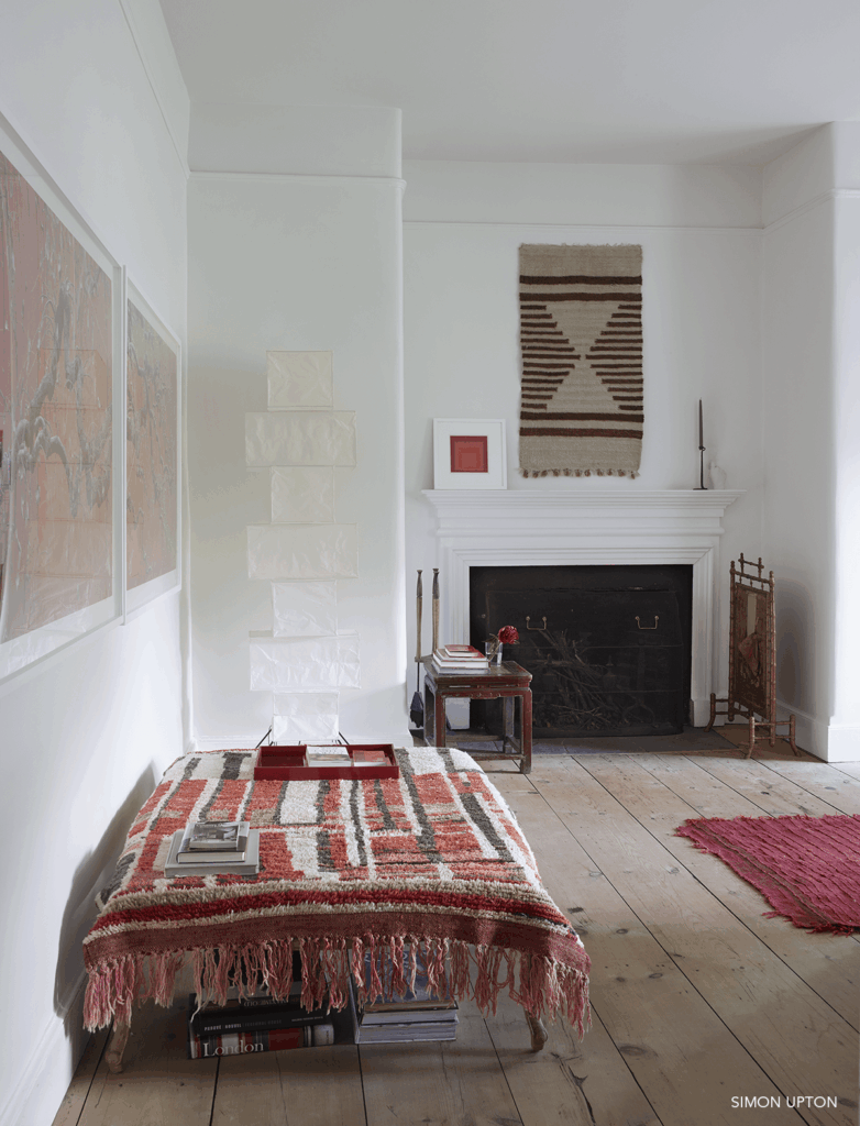Room with white walls, wood floors, a fireplace on the rear wall, a red rug and a low bench with a red patterned blanket.