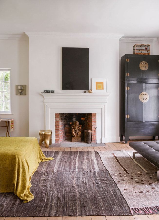 Bedroom with brick fireplace and traditional white mantel, black painting, black armoire, taupe rug, bed with yellow cover.