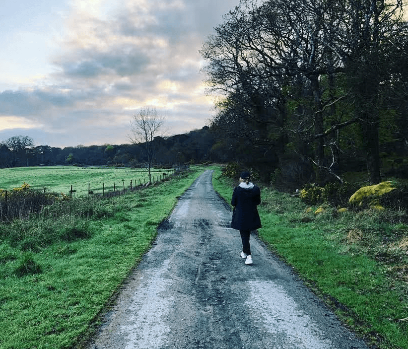 A woman in black pants, a black jacket and white sneakers walking on an old, paved road on the edge of a green countryside.