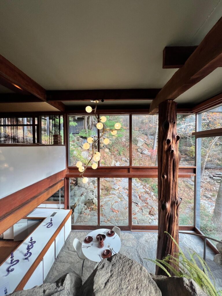 A double height interior with a wall of wood-trimmed windows, a stone floor, a hanging light fixture and a tree trunk.