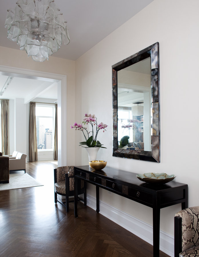 Interior with chevron pattern wood floors, white walls, black console table, pair of side chairs, mirror and glass chandelier.