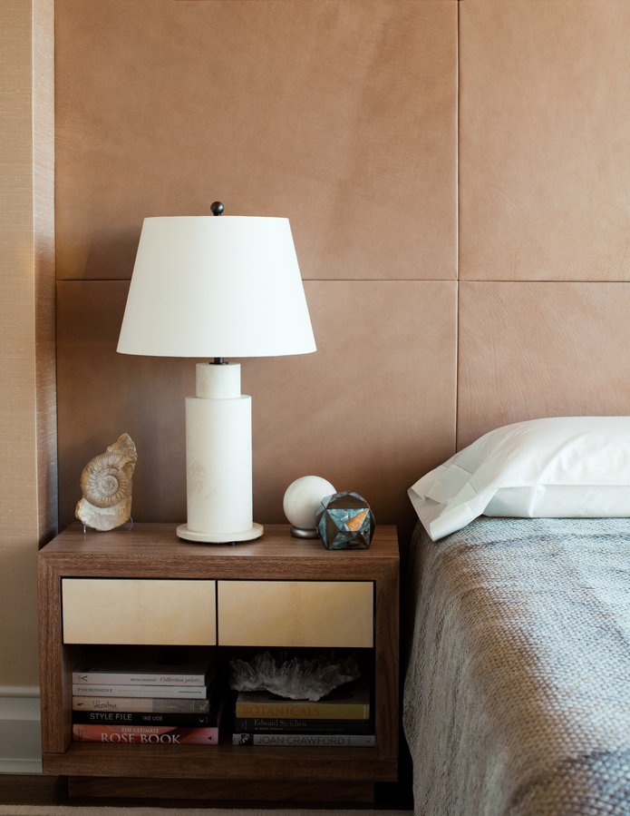 Bedside table in wood with leather drawers, modern white marble lamp, tan leather panel wall and gray chenille bedspread.