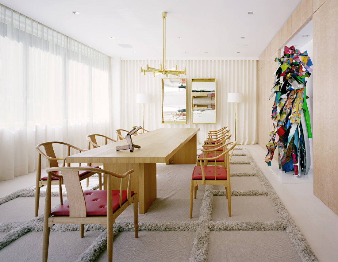 Modern dining table and chairs in oak on a beige grid-pattern carpet, two walls of white curtains, standing metal sculpture.