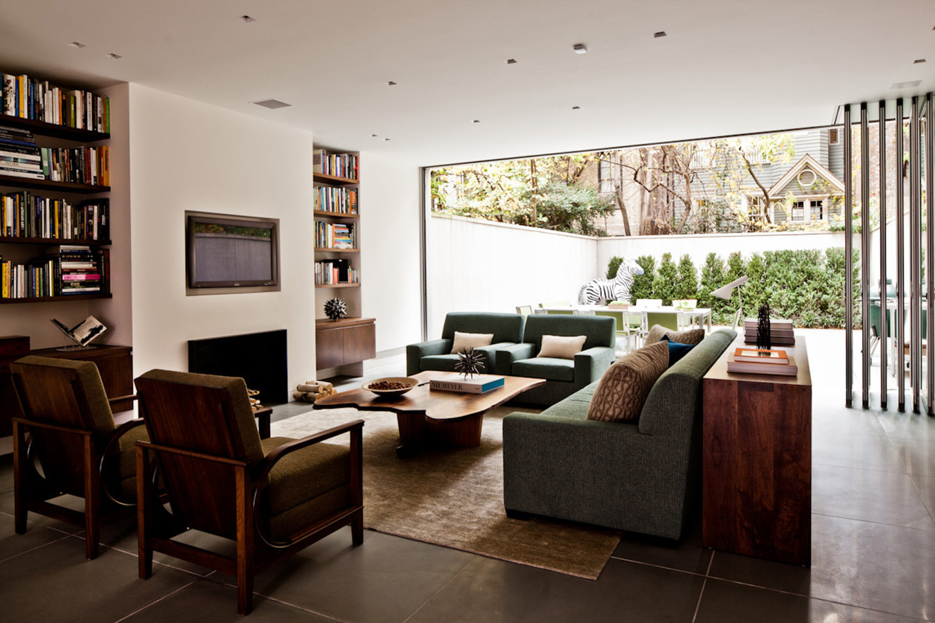 Modern living room with fireplace, bookshelves, upholstered chairs and sofa, and open back wall leading to outdoor patio.