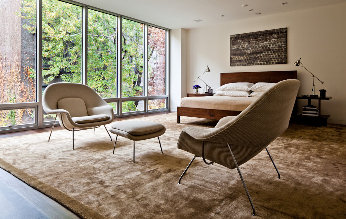 Primary bedroom with modern walnut bed and side tables, seating area and silk rug with a wall of floor-to-ceiling windows