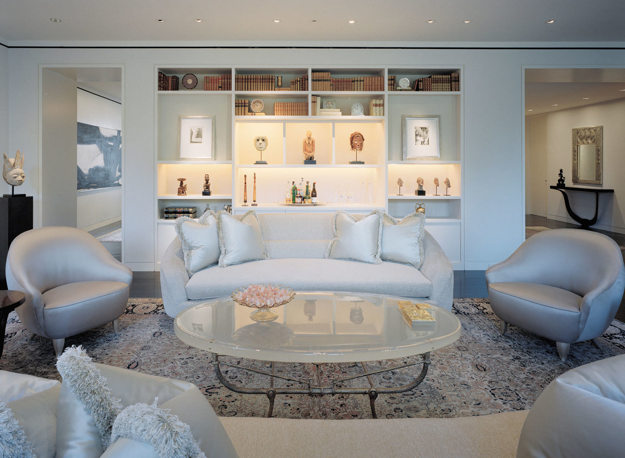 Formal living room in a white palette with upholstered seating, oval glass table and a millwork wall of books and objects.