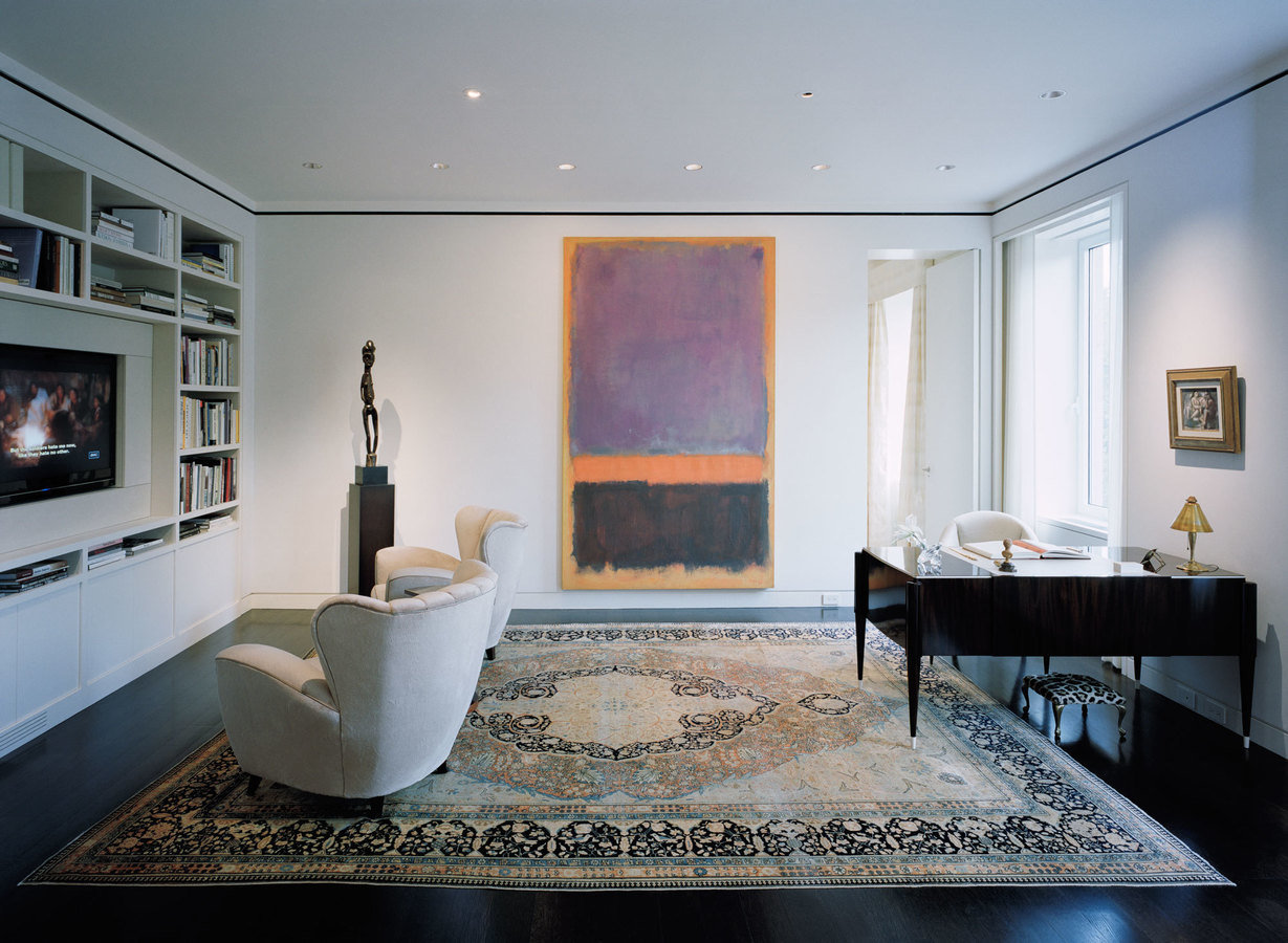 White-walled den with dark floors, antique rug, formal writing desk, pair of upholstered chairs, tv and large modern painting.