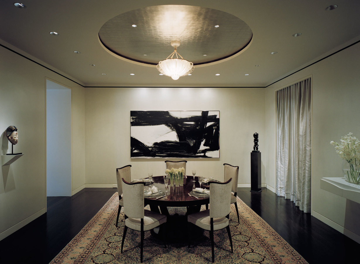 Formal dining room with circular inset ceiling and chandelier over table, dark floor, antique rug, sculptures and modern art.