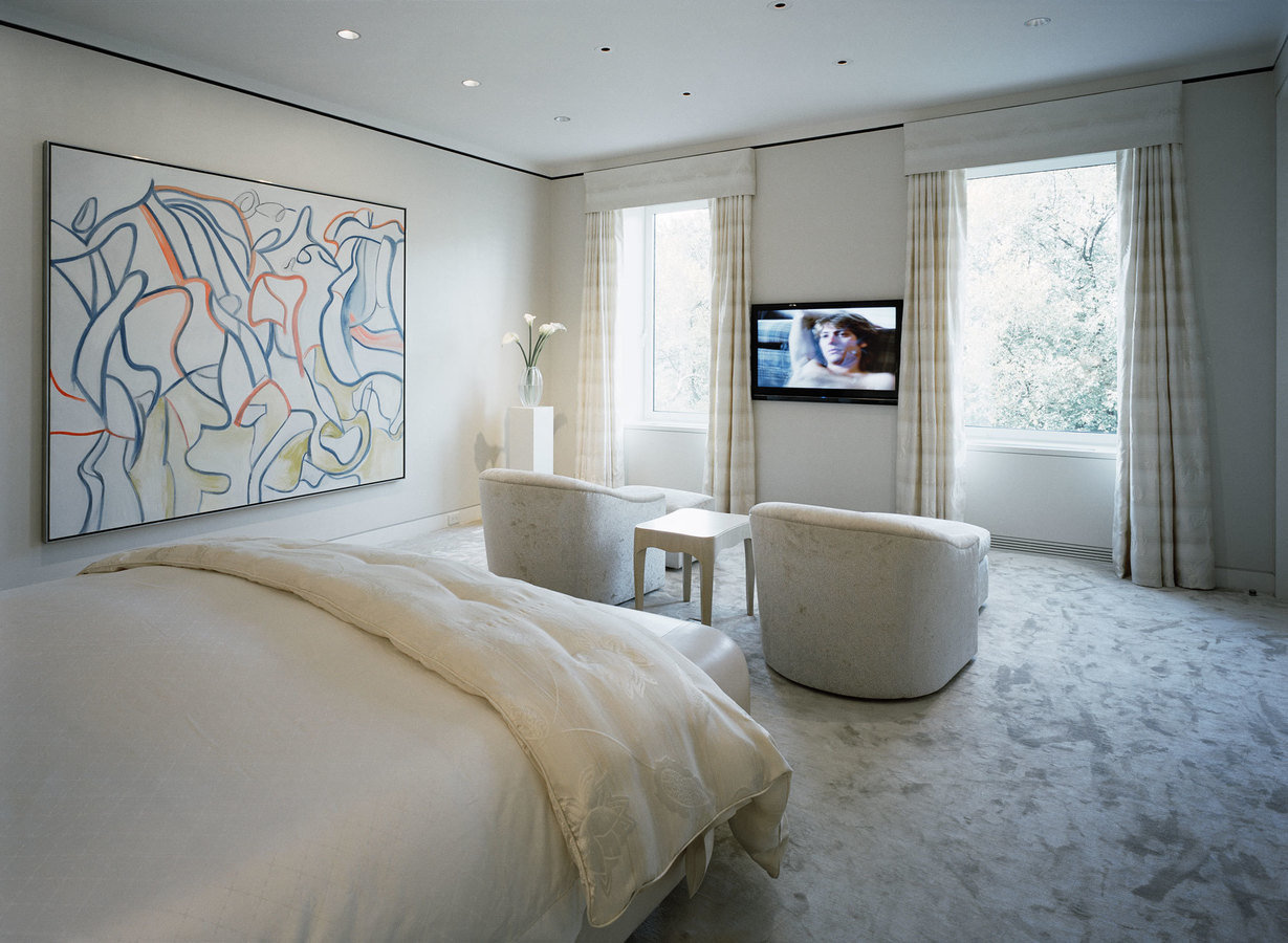 Carpeted bedroom in a white palette with two windows and tv opposite the bed, a pair of upholstered chairs and large artwork.
