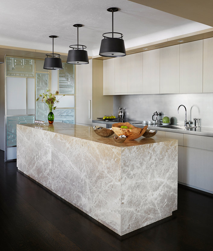 Modern kitchen in beige palette with pale cabinetry and beige marble island counter; three black suspended light fixtures.