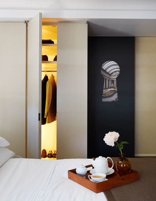 Bed in foreground with wooden tray holding a white tea set; beige wood closet door open and keyhole artwork on black wall.