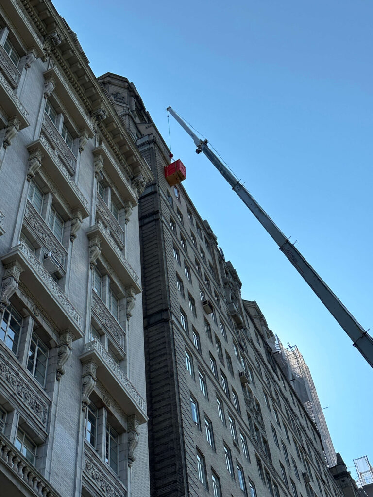 A crane hoisting a carton up to a window of a residential building with an ornate stone facade. A blue sky in the background.