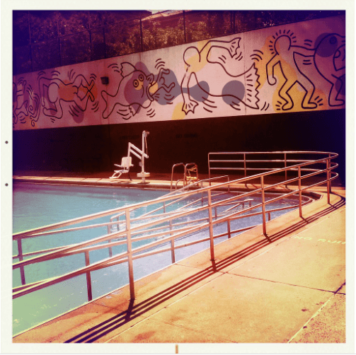 A swimming pool with a curved railing and a mural of fish and dancing figures by Keith Haring in the background.