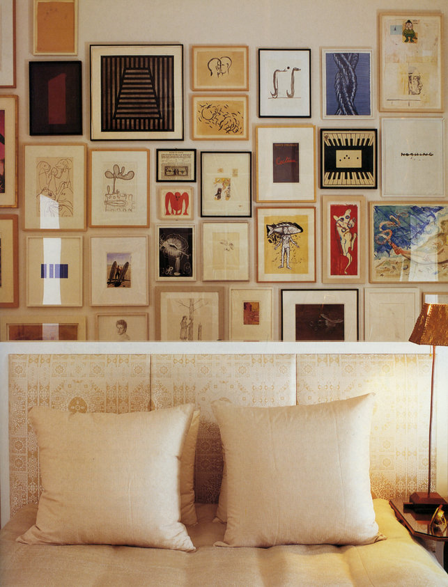 Close-up of bed with beige silk upholstered headboard, coordinating bedspread and pillows and framed art wall in background.