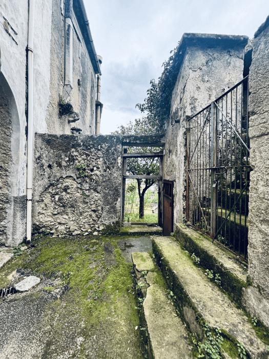 Old stone buildings with an archway on the left, stairs and a metal gate to the right and a small tree in the background.