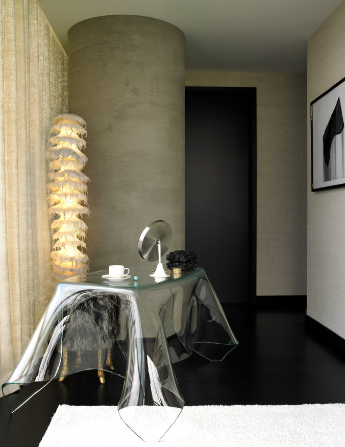 Corner of room with concrete column, black floor, white rug, silk curtains, sculptural glass table and abstract floor lamp.