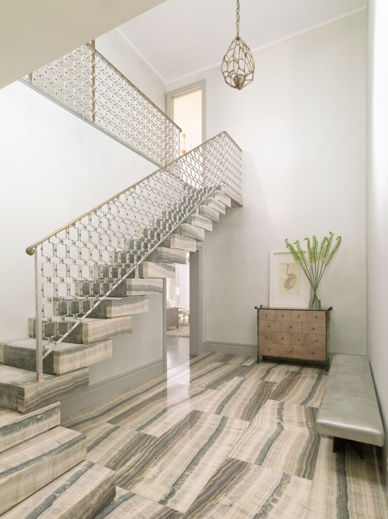 Double-height entry foyer with greige onyx stone floor and staircase with decorative metal railing and chandelier.