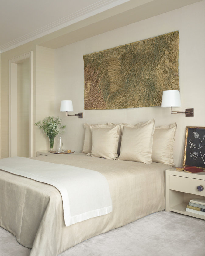 King bed with beige silk cover, off-white silk carpet, pair of sconces with white shades, end tables and gold wall-hanging.