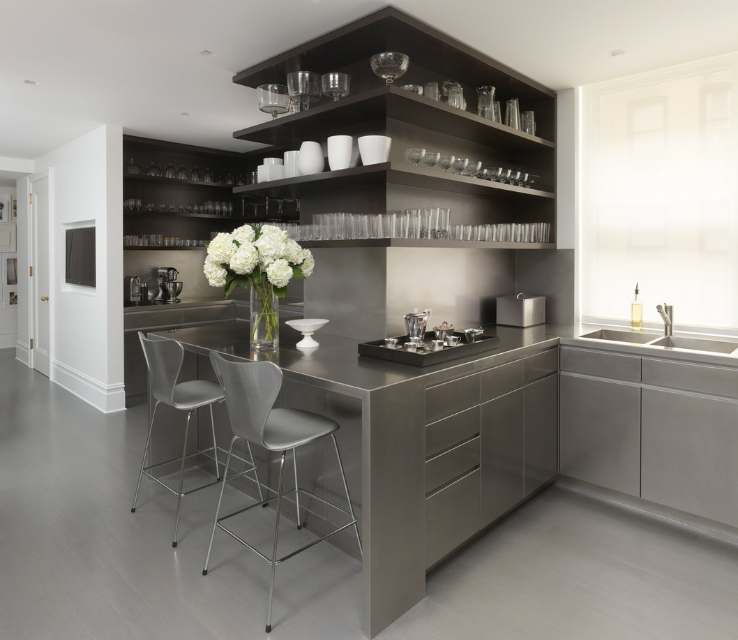 Kitchen with gray wood floor, stainless steel counter and lower cabinets with counter and stools and dark wood open shelves.